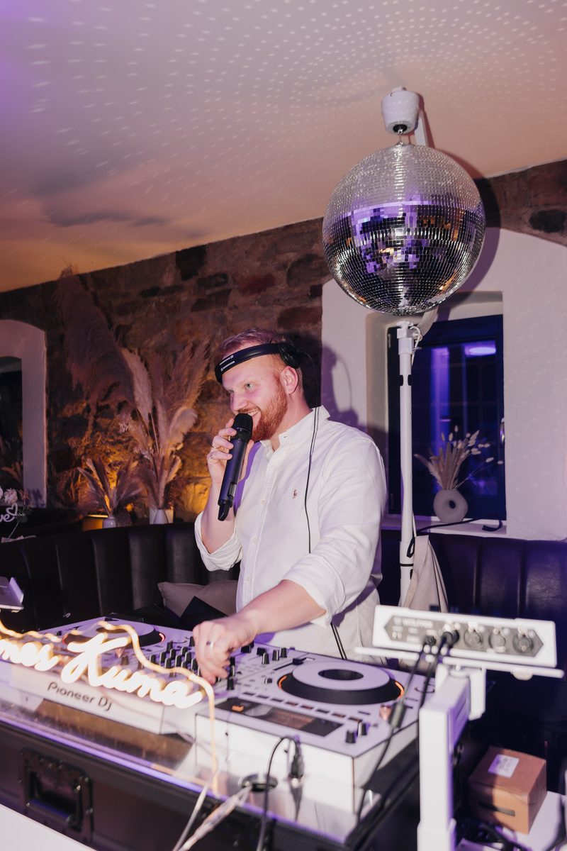James Kuma mit Mikrofon an den Decks, Discokugel über ihm