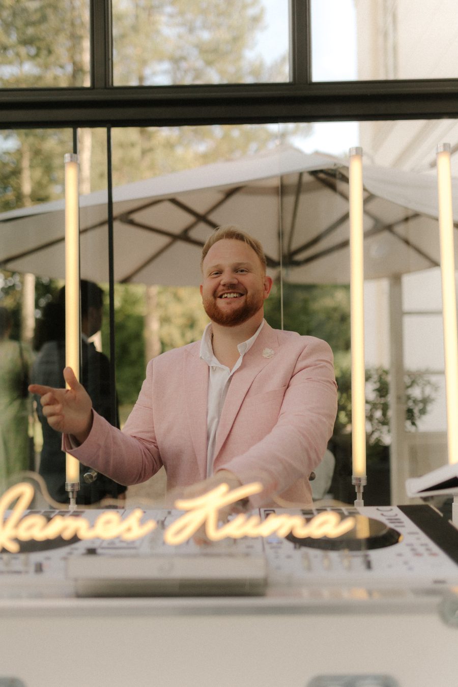 James Kuma im rosa Sakko lachend hinter der weißen Booth mit Neon-Schriftzug, warmes Nachmittagslicht auf einer Terrasse