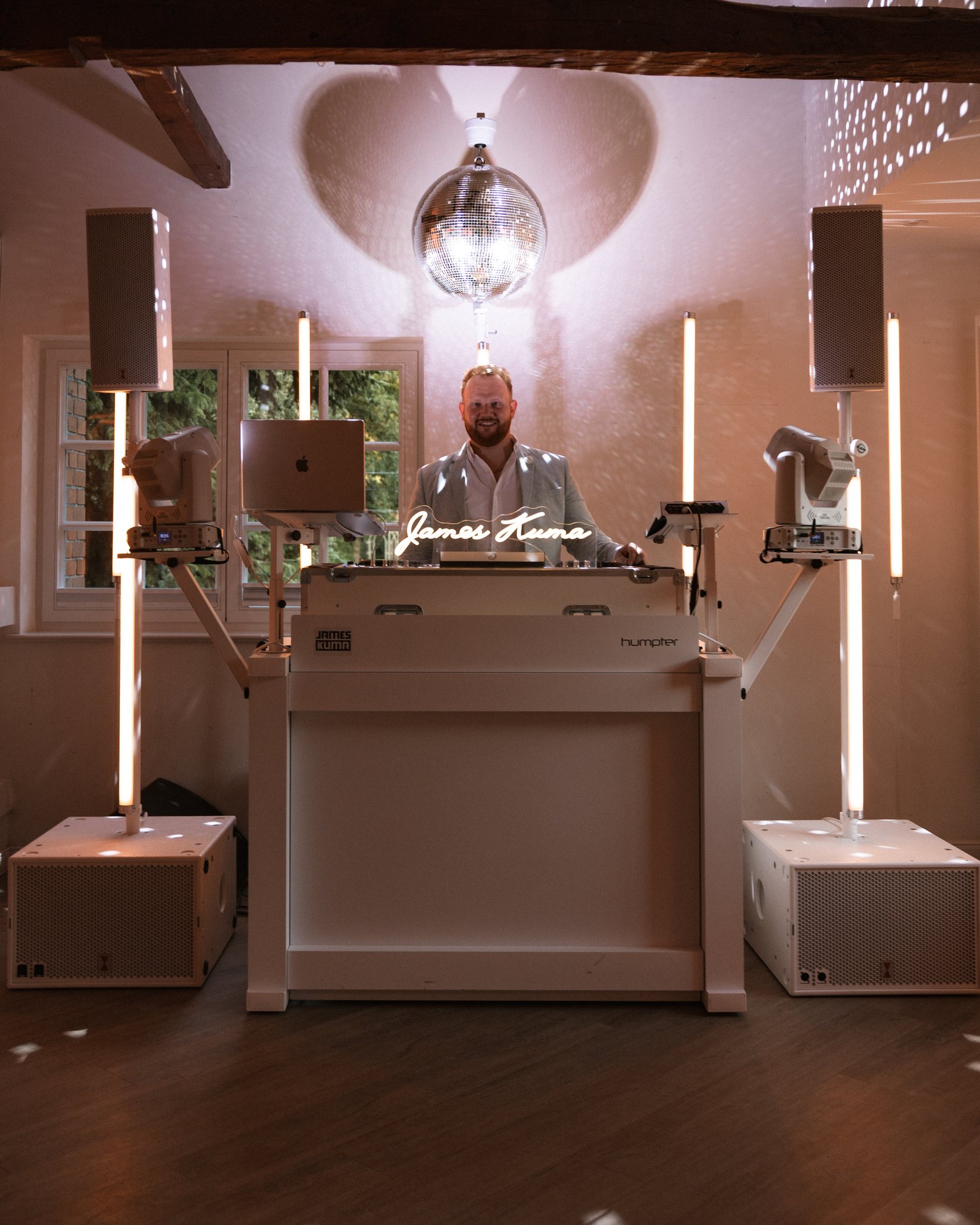 Abendlich beleuchteter Saal mit weißer DJ-Booth, Discokugel und warmen Lichtpunkten an Wand und Decke
