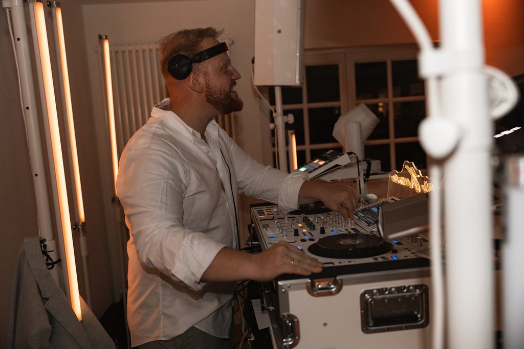 James Kuma im weißen Hemd mit Kopfhörern an den Decks, Tageslicht durch das Fenster