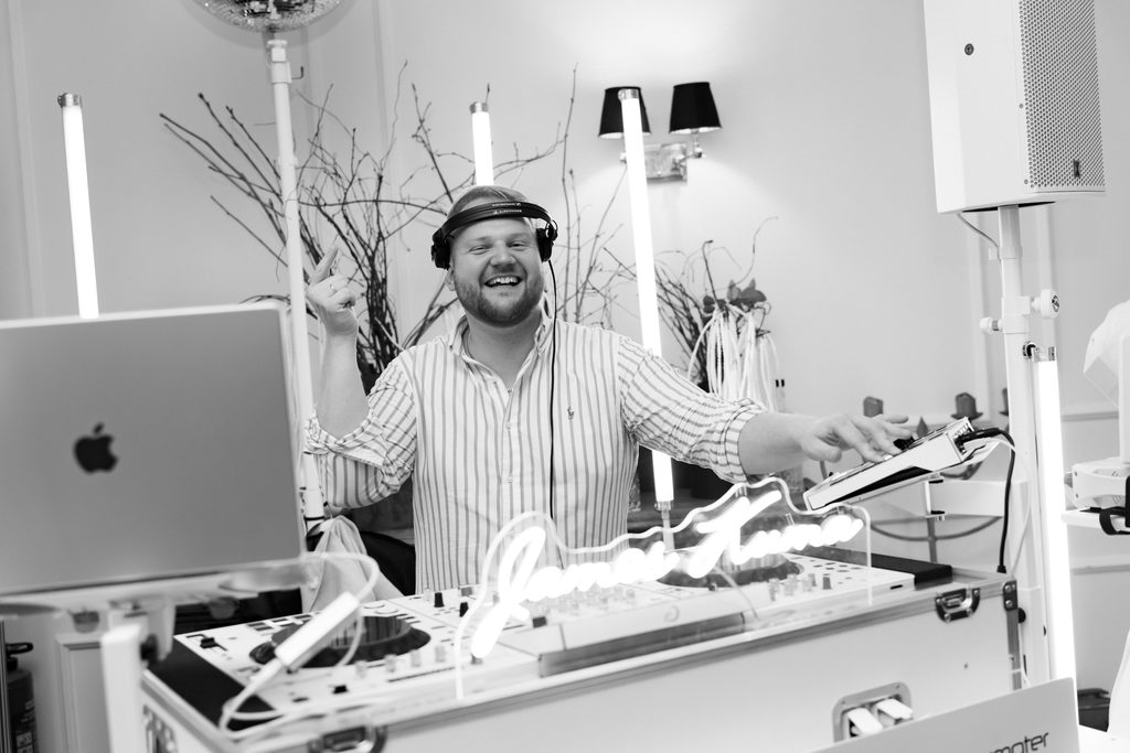 James Kuma lachend mit Kopfhörern hinter der weißen Booth, davor der Neon-Schriftzug James Kuma, weiße Lichtstelen und Discokugel, Schwarzweiß
