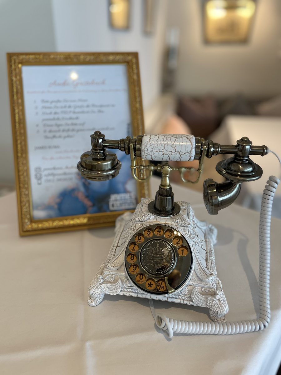 Weißes Retro-Telefon mit goldener Wählscheibe als Audio-Gästebuch, dahinter die gerahmte Anleitung