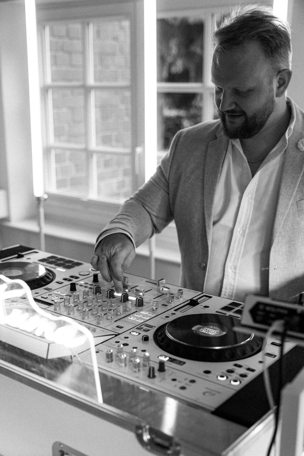 James Kuma konzentriert am Mixer, im hellen Sakko vor dem Fenster — schwarzweiß